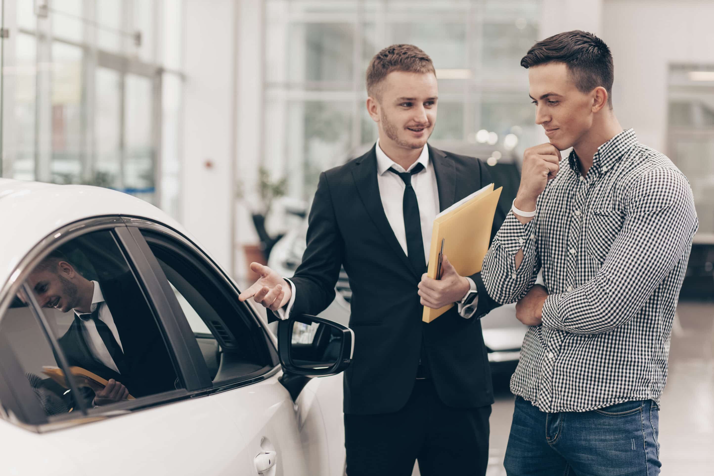 Professional car dealer showing cars on sale to his male customer, working at the auto dealership salon. Man looking thoughtfully at the car he is examining. Handsome man buying new automobile Professional car dealer showing cars on sale to his male customer, working at the auto dealership salon. Man looking thoughtfully at the car he is examining. Handsome man buying new automobile with auto loan
