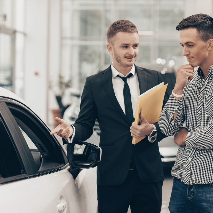 Untitled design (2) Professional car dealer showing cars on sale to his male customer, working at the auto dealership salon. Man looking thoughtfully at the car he is examining. Handsome man buying new automobile with auto loan